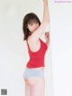 A woman in a red tank top and blue panties leaning against a wall.